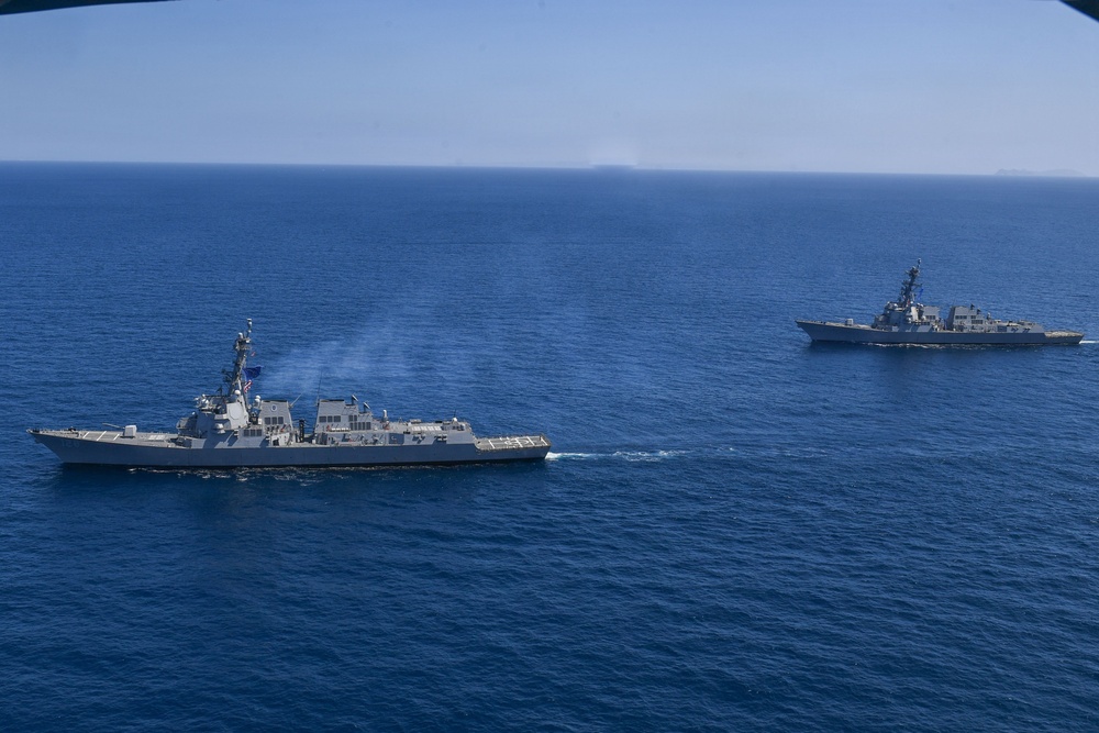 An aerial photograph shows two naval warships underway in open blue ocean waters. Both vessels appear to be guided-missile destroyers (DDG-class) traveling in formation, with the closer ship emitting light exhaust smoke from its stack. The image was likely taken from a helicopter or aircraft, providing an elevated perspective of the maritime operation.