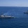 Pre-Commissioning Unit Ted Stevens, USS Daniel Inouye sail together An aerial photograph shows two naval warships underway in open blue ocean waters. Both vessels appear to be guided-missile destroyers (DDG-class) traveling in formation, with the closer ship emitting light exhaust smoke from its stack. The image was likely taken from a helicopter or aircraft, providing an elevated perspective of the maritime operation.