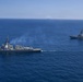 An aerial photograph shows two naval warships underway in open blue ocean waters. Both vessels appear to be guided-missile destroyers (DDG-class) traveling in formation, with the closer ship emitting light exhaust smoke from its stack. The image was likely taken from a helicopter or aircraft, providing an elevated perspective of the maritime operation.