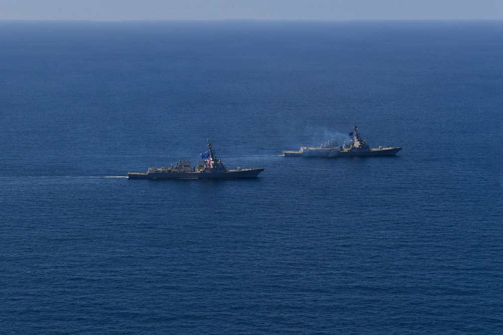 Two naval warships, appearing to be Arleigh Burke-class guided-missile destroyers of the United States Navy, are underway in open blue ocean waters. Both vessels are sailing in a parallel formation, with one ship visibly emitting smoke or exhaust from its superstructure. An American flag and at least one additional flag are visible on the nearer vessel.