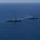Pre-Commissioning Unit Ted Stevens, USS Daniel Inouye sail together Two naval warships, appearing to be Arleigh Burke-class guided-missile destroyers of the United States Navy, are underway in open blue ocean waters. Both vessels are sailing in a parallel formation, with one ship visibly emitting smoke or exhaust from its superstructure. An American flag and at least one additional flag are visible on the nearer vessel.