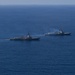 Two naval warships, appearing to be Arleigh Burke-class guided-missile destroyers of the United States Navy, are underway in open blue ocean waters. Both vessels are sailing in a parallel formation, with one ship visibly emitting smoke or exhaust from its superstructure. An American flag and at least one additional flag are visible on the nearer vessel.