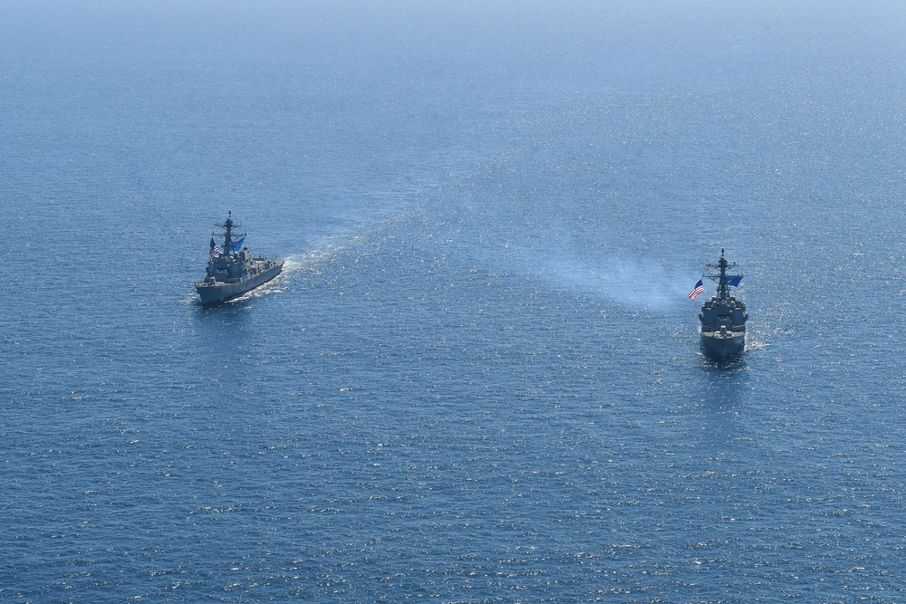 Two naval warships are underway in open blue ocean waters, photographed from an elevated aerial perspective. Both vessels are flying American flags and appear to be operating in formation or conducting a joint naval exercise. One ship on the right appears to be emitting exhaust smoke from its propulsion system.