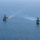 Pre-Commissioning Unit Ted Stevens, USS Daniel Inouye sail together Two naval warships are underway in open blue ocean waters, photographed from an elevated aerial perspective. Both vessels are flying American flags and appear to be operating in formation or conducting a joint naval exercise. One ship on the right appears to be emitting exhaust smoke from its propulsion system.
