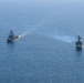 Two naval warships are underway in open blue ocean waters, photographed from an elevated aerial perspective. Both vessels are flying American flags and appear to be operating in formation or conducting a joint naval exercise. One ship on the right appears to be emitting exhaust smoke from its propulsion system.