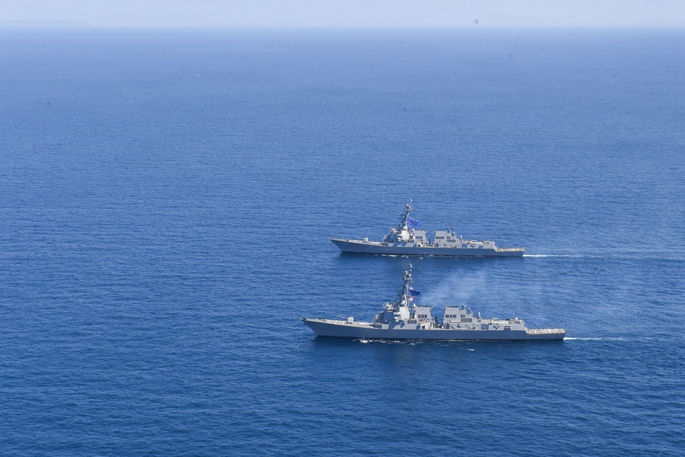 Two gray naval warships are sailing in formation across open blue ocean waters. The foreground vessel appears to be emitting smoke or exhaust from its superstructure, suggesting active propulsion or weapons activity. Both vessels appear to be modern guided-missile destroyers or frigates based on their hull profiles and superstructure configurations.