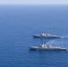 Two gray naval warships are sailing in formation across open blue ocean waters. The foreground vessel appears to be emitting smoke or exhaust from its superstructure, suggesting active propulsion or weapons activity. Both vessels appear to be modern guided-missile destroyers or frigates based on their hull profiles and superstructure configurations.