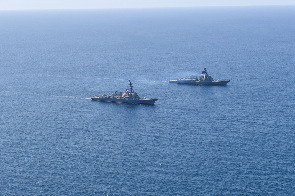 An aerial photograph shows two naval warships sailing in formation across open blue ocean waters. The foreground vessel displays a visible American flag, identifying it as a US Navy warship, likely an Arleigh Burke-class guided-missile destroyer. Both ships appear to be underway at moderate speed, leaving visible wakes in calm sea conditions.
