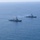 Pre-Commissioning Unit Ted Stevens, USS Daniel Inouye sail together An aerial photograph shows two naval warships sailing in formation across open blue ocean waters. The foreground vessel displays a visible American flag, identifying it as a US Navy warship, likely an Arleigh Burke-class guided-missile destroyer. Both ships appear to be underway at moderate speed, leaving visible wakes in calm sea conditions.