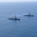 An aerial photograph shows two naval warships sailing in formation across open blue ocean waters. The foreground vessel displays a visible American flag, identifying it as a US Navy warship, likely an Arleigh Burke-class guided-missile destroyer. Both ships appear to be underway at moderate speed, leaving visible wakes in calm sea conditions.