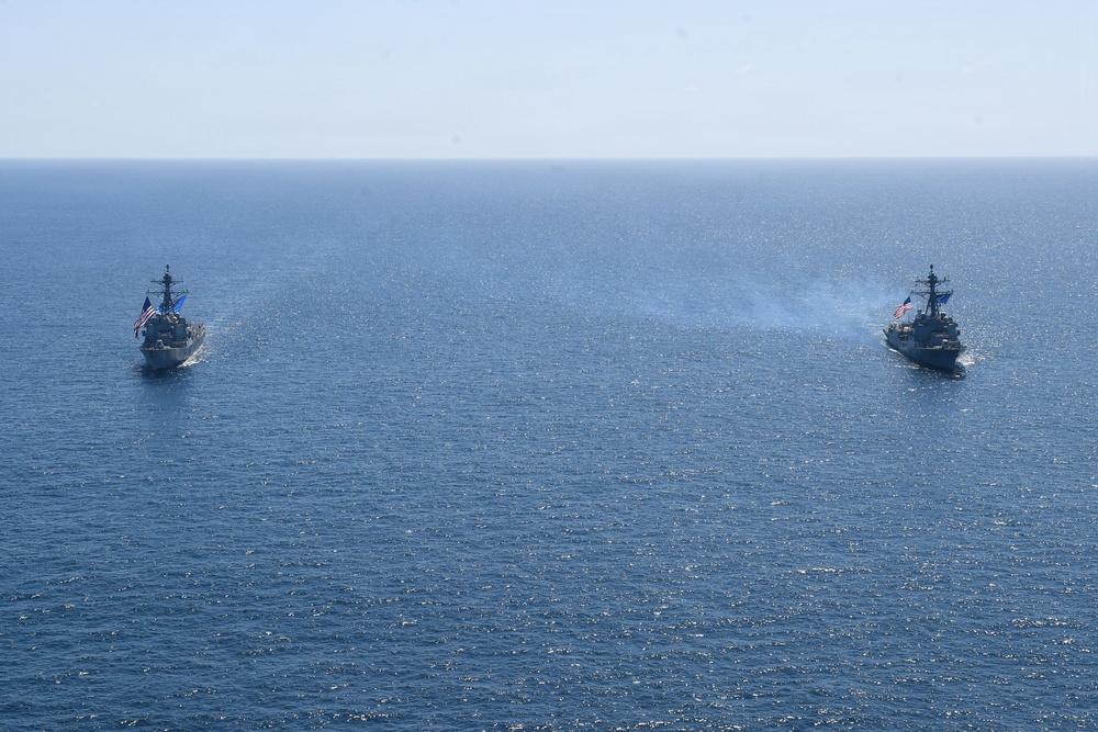Two U.S. Navy warships are underway in open blue ocean, sailing in parallel formation. Both vessels are flying American flags and appear to be Arleigh Burke-class guided-missile destroyers. The ship on the right is emitting light smoke or exhaust, suggesting active propulsion or a recent firing event.