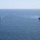 Pre-Commissioning Unit Ted Stevens, USS Daniel Inouye sail together Two U.S. Navy warships are underway in open blue ocean, sailing in parallel formation. Both vessels are flying American flags and appear to be Arleigh Burke-class guided-missile destroyers. The ship on the right is emitting light smoke or exhaust, suggesting active propulsion or a recent firing event.