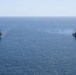 Two U.S. Navy warships are underway in open blue ocean, sailing in parallel formation. Both vessels are flying American flags and appear to be Arleigh Burke-class guided-missile destroyers. The ship on the right is emitting light smoke or exhaust, suggesting active propulsion or a recent firing event.