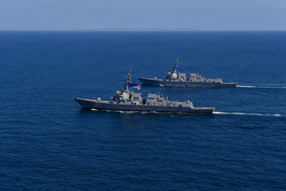 Two gray naval warships, consistent with Arleigh Burke-class guided-missile destroyers, are sailing in formation across open blue ocean waters. The foreground vessel displays a United States flag, and both ships appear to be underway at moderate speed, leaving visible wakes. The image appears to document a naval formation exercise or transit operation.