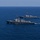 Pre-Commissioning Unit Ted Stevens, USS Daniel Inouye sail together Two gray naval warships, consistent with Arleigh Burke-class guided-missile destroyers, are sailing in formation across open blue ocean waters. The foreground vessel displays a United States flag, and both ships appear to be underway at moderate speed, leaving visible wakes. The image appears to document a naval formation exercise or transit operation.