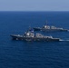 Two gray naval warships, consistent with Arleigh Burke-class guided-missile destroyers, are sailing in formation across open blue ocean waters. The foreground vessel displays a United States flag, and both ships appear to be underway at moderate speed, leaving visible wakes. The image appears to document a naval formation exercise or transit operation.