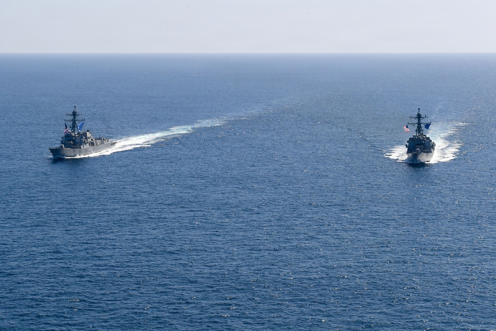 Two U.S. Navy warships are sailing in formation across open blue ocean waters. Both vessels appear to be Arleigh Burke-class guided-missile destroyers, identifiable by their gray hulls, superstructure configuration, and American flags. The ships are traveling on parallel courses with visible white wake trails behind them.