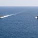 Two U.S. Navy warships are sailing in formation across open blue ocean waters. Both vessels appear to be Arleigh Burke-class guided-missile destroyers, identifiable by their gray hulls, superstructure configuration, and American flags. The ships are traveling on parallel courses with visible white wake trails behind them.