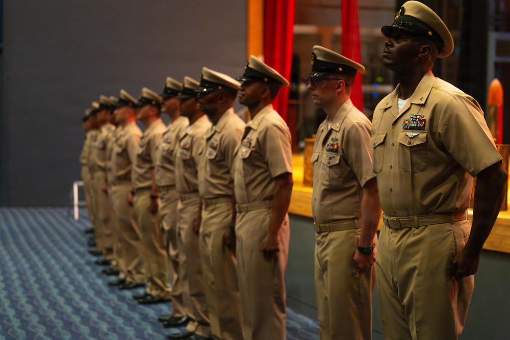 A formation of U.S. Navy personnel in Service Khaki uniforms (Type B) standing at attention during what appears to be a formal military ceremony. Approximately 10-12 sailors are lined up in a diagonal formation, with the foreground figure displaying multiple service ribbons and warfare qualification badges. The setting appears to be an indoor venue with a stage or platform visible in the background.