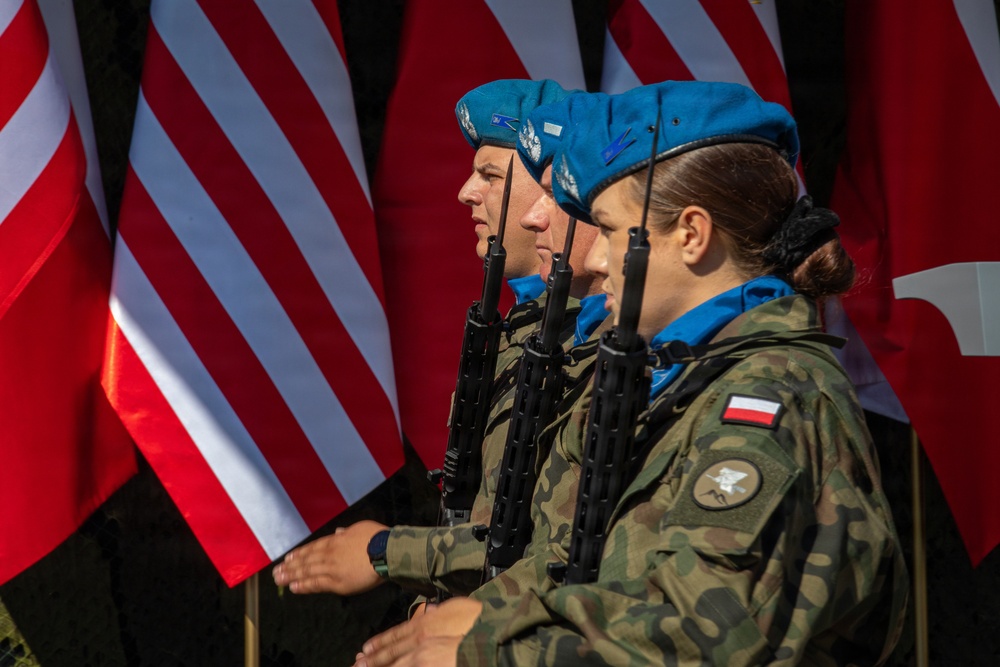 Three military personnel wearing blue berets and camouflage uniforms stand at attention in a formal ceremony. They are carrying rifles and wearing patches identifying them as Polish military, with blue scarves indicating a UN peacekeeping affiliation. Multiple flags, including the United States flag and what appears to be the Polish flag, are visible in the background.