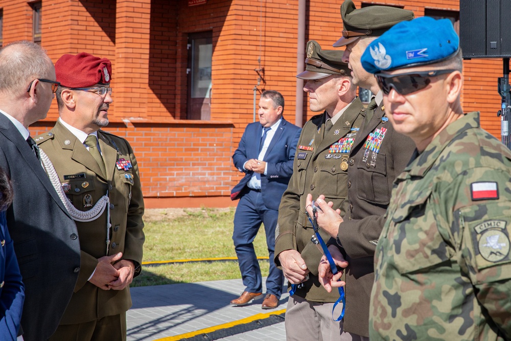 A group of military personnel from multiple nations are engaged in conversation at what appears to be an outdoor military ceremony or official event. The scene includes soldiers wearing Polish Army uniforms with distinctive berets (red and blue), a US Army officer with extensive medal ribbons, and a civilian in a suit standing in the background. The setting is in front of a red brick building, suggesting a military installation.