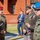 Oleszno cultural center dedicated to Staff Sgt. Michael Ollis, honoring U.S.-Poland bond A group of military personnel from multiple nations are engaged in conversation at what appears to be an outdoor military ceremony or official event. The scene includes soldiers wearing Polish Army uniforms with distinctive berets (red and blue), a US Army officer with extensive medal ribbons, and a civilian in a suit standing in the background. The setting is in front of a red brick building, suggesting a military installation.