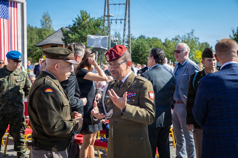 A military ceremony or official event appears to be taking place outdoors, featuring both American and Polish military personnel in conversation. Two senior officers are prominently engaged in discussion — one wearing a US Army uniform with a campaign hat, and another in a Polish Army uniform with a distinctive maroon beret bearing four stars and the Polish Eagle insignia. An American flag is visible in the background alongside a crowd of military and civilian attendees.