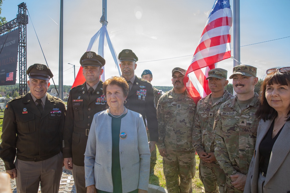 A group of U.S. military personnel in various uniforms pose alongside civilian officials in front of raised American and Polish flags. The setting appears to be an outdoor ceremony, likely at a military installation or public venue in Poland. Name tapes visible on some soldiers include 'BAYLESS' and 'BRASHER'.