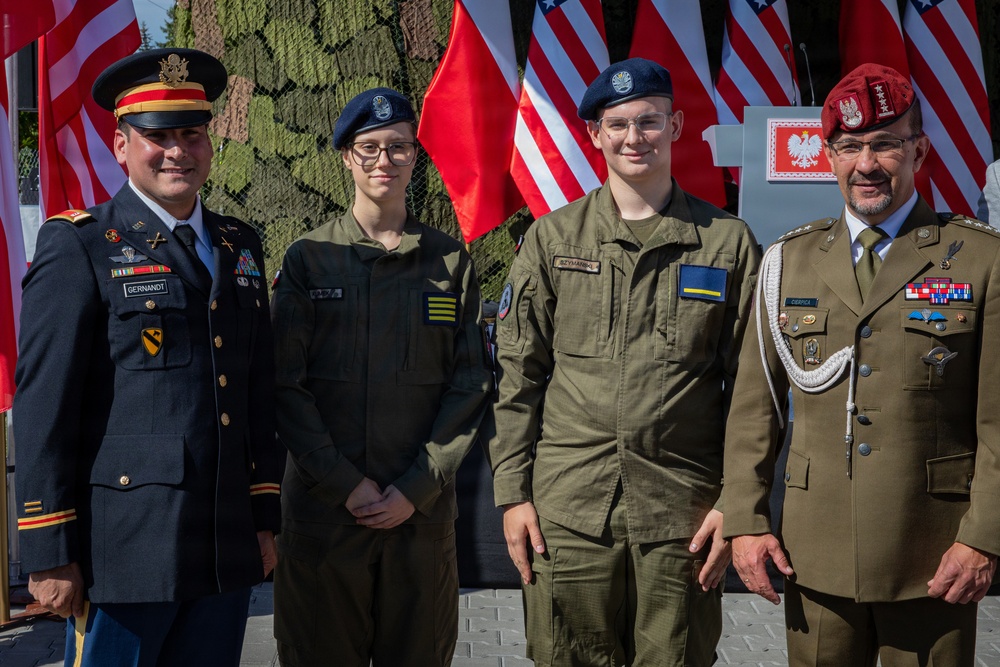 Four individuals in military uniforms pose together at what appears to be a joint US-Polish military ceremony or event. Two appear to be senior officers (one US Army, one Polish Army), and two appear to be younger cadets or junior personnel wearing blue berets. American and Polish flags are prominently displayed in the background.