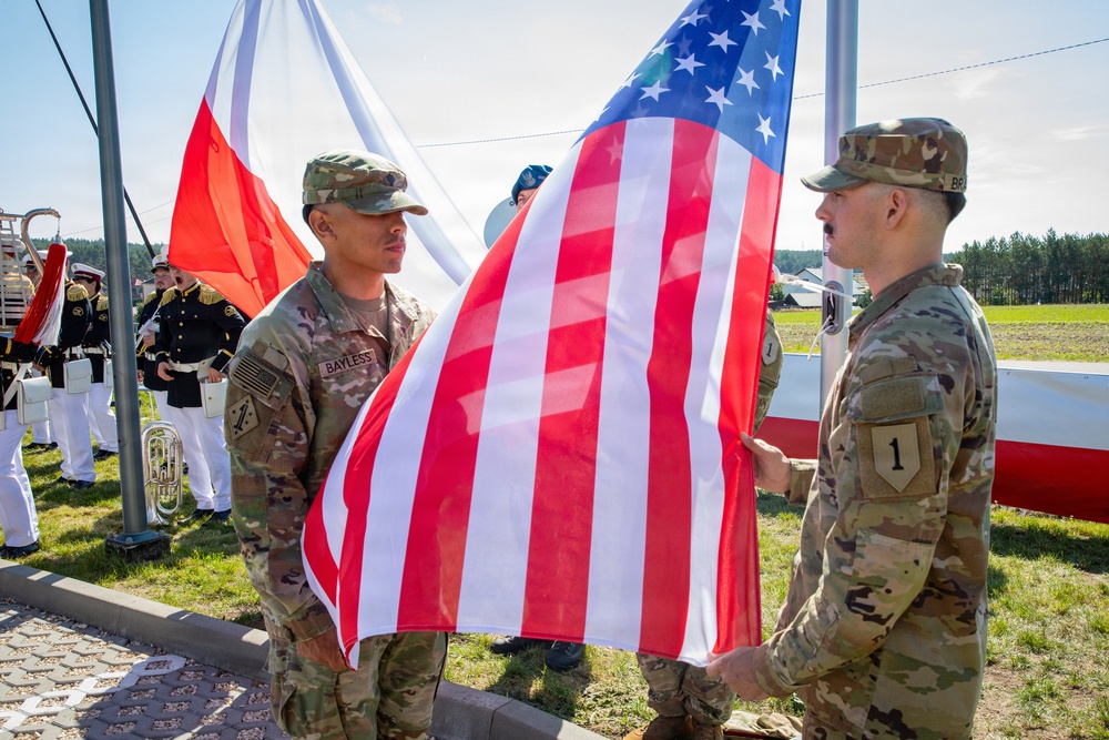 Two U.S. Army soldiers in MultiCam/OCP camouflage uniforms are holding and preparing to raise an American flag during a formal military ceremony. A Polish flag is visible in the background, along with a military band in dress uniforms, suggesting a bilateral or NATO-related event likely held in Poland. The soldier on the left is identified by his name tape as 'BAYLESS,' and the soldier on the right wears a '1st Infantry Division' shoulder patch.