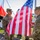 Oleszno cultural center dedicated to Staff Sgt. Michael Ollis, honoring U.S.-Poland bond Two U.S. Army soldiers in MultiCam/OCP camouflage uniforms are holding and preparing to raise an American flag during a formal military ceremony. A Polish flag is visible in the background, along with a military band in dress uniforms, suggesting a bilateral or NATO-related event likely held in Poland. The soldier on the left is identified by his name tape as 'BAYLESS,' and the soldier on the right wears a '1st Infantry Division' shoulder patch.