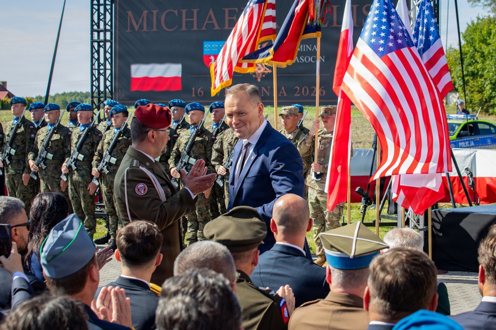 A formal military ceremony is taking place outdoors, likely in Poland, featuring both Polish and American military personnel alongside civilian dignitaries. A man in a blue suit is shaking hands with a military officer wearing a red beret, while rows of soldiers stand at attention in the background. Multiple American and Polish flags are displayed prominently, and a banner dated '16 [CZERW]CA 2026' is visible in the background.