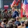 A formal military ceremony is taking place outdoors, likely in Poland, featuring both Polish and American military personnel alongside civilian dignitaries. A man in a blue suit is shaking hands with a military officer wearing a red beret, while rows of soldiers stand at attention in the background. Multiple American and Polish flags are displayed prominently, and a banner dated '16 [CZERW]CA 2026' is visible in the background.