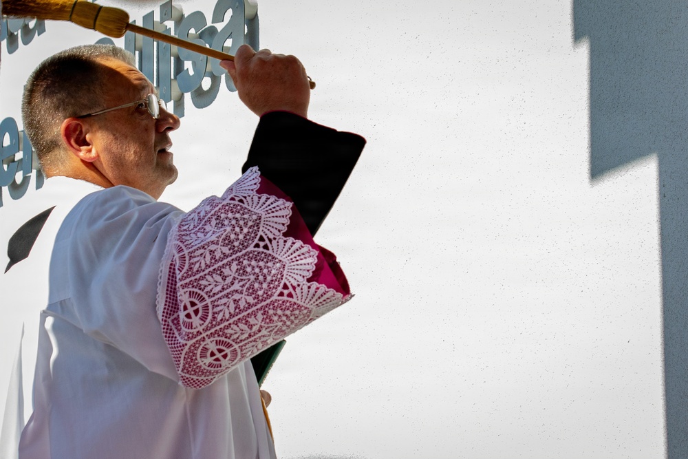 A Catholic clergy member, dressed in a white alb with ornate lace-trimmed vestments and a magenta/pink stole, is performing a blessing ritual outdoors. He is holding an aspergillum (holy water sprinkler) raised upward in one hand and a black-covered book (likely a ritual or liturgical book) in the other. The setting appears to be an outdoor religious ceremony, possibly a blessing or dedication of a building.