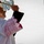 Oleszno cultural center dedicated to Staff Sgt. Michael Ollis, honoring U.S.-Poland bond A Catholic clergy member, dressed in a white alb with ornate lace-trimmed vestments and a magenta/pink stole, is performing a blessing ritual outdoors. He is holding an aspergillum (holy water sprinkler) raised upward in one hand and a black-covered book (likely a ritual or liturgical book) in the other. The setting appears to be an outdoor religious ceremony, possibly a blessing or dedication of a building.