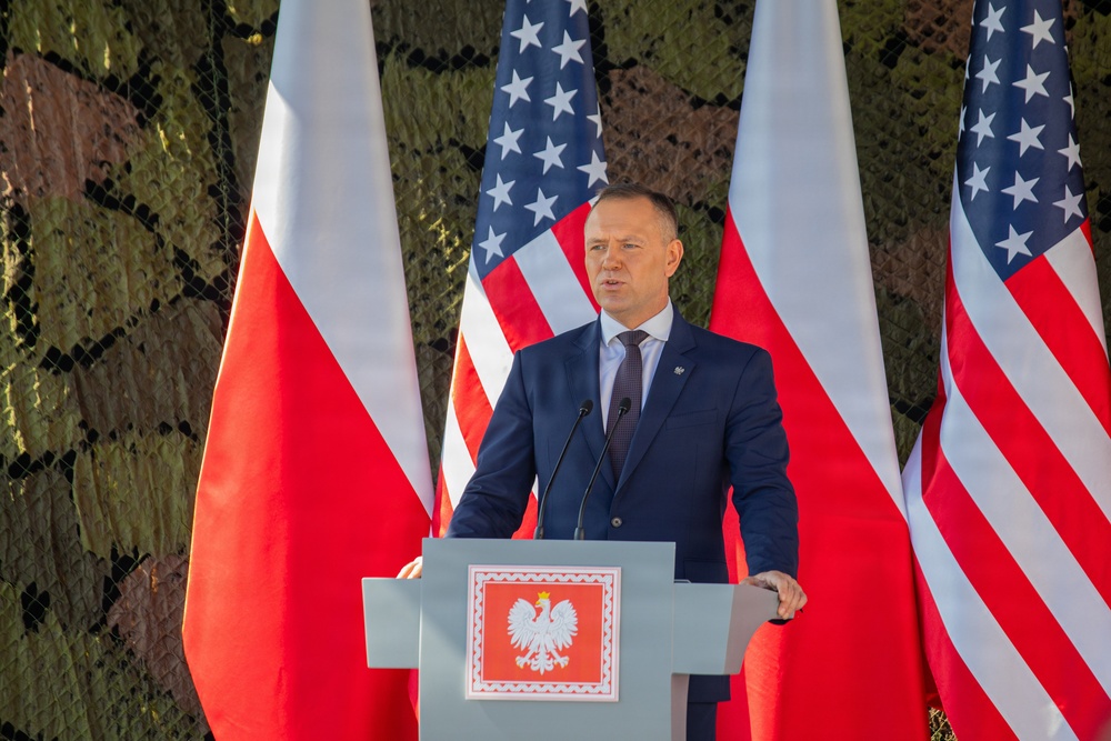 A man in a dark navy suit is speaking at a podium bearing the Polish coat of arms (white eagle on red background). The setting appears to be an official bilateral event between Poland and the United States, as evidenced by alternating Polish and American flags displayed prominently behind the speaker. The background features military-style camouflage netting, suggesting a defense or military-related ceremony.