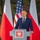 Oleszno cultural center dedicated to Staff Sgt. Michael Ollis, honoring U.S.-Poland bond A man in a dark navy suit is speaking at a podium bearing the Polish coat of arms (white eagle on red background). The setting appears to be an official bilateral event between Poland and the United States, as evidenced by alternating Polish and American flags displayed prominently behind the speaker. The background features military-style camouflage netting, suggesting a defense or military-related ceremony.