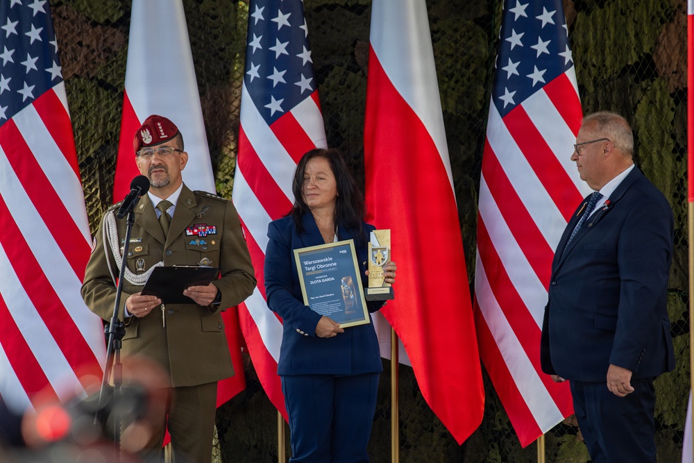 A formal award ceremony is taking place outdoors with three individuals standing before alternating Polish and American flags. A Polish military officer in uniform speaks at a microphone while a woman in a blue suit holds a certificate and trophy. The event appears to be the 'Warszawskie Targi Obronne' (Warsaw Defence Exhibition) award presentation.