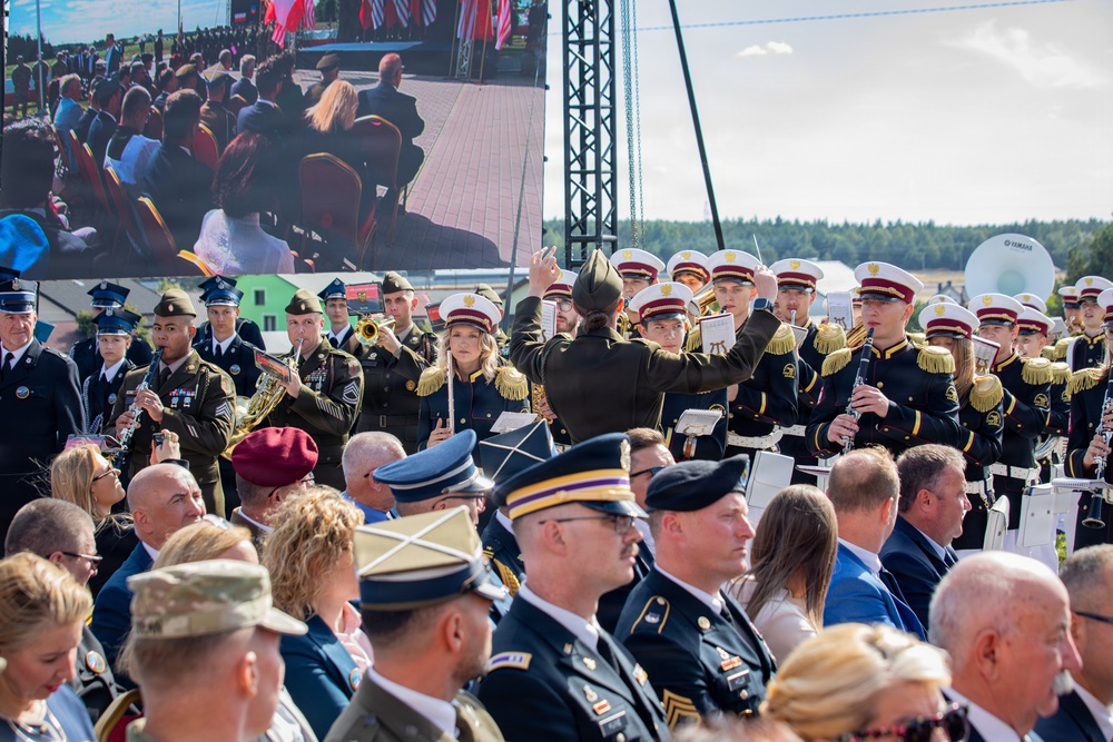 A formal military ceremony featuring a joint military band performance with musicians in multiple uniform types, including dark dress uniforms with white caps and gold epaulettes, and standard U.S. Army service uniforms. A conductor in a dark uniform with a distinctive hat is visible from behind, directing the ensemble. A large LED screen in the background displays a live feed of the seated audience, which includes military officers and civilians.