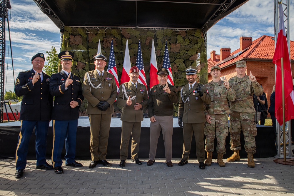 A group of eight military personnel pose together in front of a stage decorated with American and Polish flags, suggesting a joint US-Poland military ceremony or event. The individuals wear a mix of uniforms including US Army dress blues, US Army combat uniforms (ACU/OCP), and Polish military dress uniforms. Several individuals are making a 'number one' hand gesture with their index fingers raised.