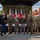 Oleszno cultural center dedicated to Staff Sgt. Michael Ollis, honoring U.S.-Poland bond A group of eight military personnel pose together in front of a stage decorated with American and Polish flags, suggesting a joint US-Poland military ceremony or event. The individuals wear a mix of uniforms including US Army dress blues, US Army combat uniforms (ACU/OCP), and Polish military dress uniforms. Several individuals are making a 'number one' hand gesture with their index fingers raised.