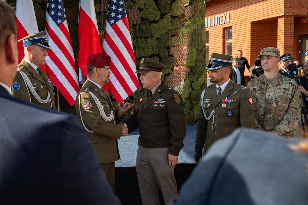 A formal military ceremony is taking place outdoors, featuring personnel from both the United States Army and the Polish Armed Forces. Two senior officers are engaged in a handshake, suggesting an official greeting, award presentation, or bilateral military engagement. American and Polish flags are prominently displayed in the background, indicating a joint or allied military event.