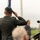 Cpt. Dolf Janssen Salutes during the U.S. National Anthem, Operation Market Garden Commemorations 2026 A military officer in dress uniform is performing a formal salute at what appears to be an outdoor ceremony or commemoration event. The officer is photographed from behind, standing at a podium with a microphone, wearing a dark green beret and uniform with decorative shoulder cord. A flagpole and small audience are visible in the background.