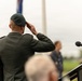 A military officer in dress uniform is performing a formal salute at what appears to be an outdoor ceremony or commemoration event. The officer is photographed from behind, standing at a podium with a microphone, wearing a dark green beret and uniform with decorative shoulder cord. A flagpole and small audience are visible in the background.
