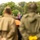 Deputy Mayor of Grave Delivers Remarks, Operation Market Garden Commemorations 2026 An official ceremony is taking place outdoors, with a man wearing a mayoral chain speaking at a podium. The audience includes individuals dressed in WWII-era military uniforms, suggesting a commemorative or liberation event. The foreground is intentionally blurred with two figures in olive-green jackets framing the scene.