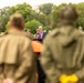 An official ceremony is taking place outdoors, with a man wearing a mayoral chain speaking at a podium. The audience includes individuals dressed in WWII-era military uniforms, suggesting a commemorative or liberation event. The foreground is intentionally blurred with two figures in olive-green jackets framing the scene.