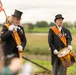 Two elderly men dressed in formal ceremonial attire — black tailcoats, top hats, and orange sashes — participate in what appears to be a Dutch civic or historical commemoration event outdoors. One man carries a heraldic flag on a pole while the other holds a decorated drum with drumsticks. The setting is a rural or semi-rural landscape with green fields visible in the background.