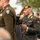 Attendees Salute at the LT John S Thompson Bridge Ceremony, Operation Market Garden Commemorations 2026 A military ceremony featuring soldiers in dress uniforms rendering salutes. The central figure is a soldier wearing a dark beret with an orange patch, holding a ceremonial baton or swagger stick, and displaying multiple unit insignia including a parachute badge and an orange-and-blue aiguillette. The background shows a crowd of civilians and other military personnel in various uniforms.