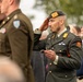 A military ceremony featuring soldiers in dress uniforms rendering salutes. The central figure is a soldier wearing a dark beret with an orange patch, holding a ceremonial baton or swagger stick, and displaying multiple unit insignia including a parachute badge and an orange-and-blue aiguillette. The background shows a crowd of civilians and other military personnel in various uniforms.