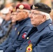 The image shows elderly veterans seated at what appears to be a commemorative or remembrance ceremony. Two men in the foreground wear dark blue Legion uniforms with Canadian Royal Canadian Legion badges and berets — one maroon/burgundy and one black. The setting appears to be an outdoor public event with a crowd visible in the blurred background.